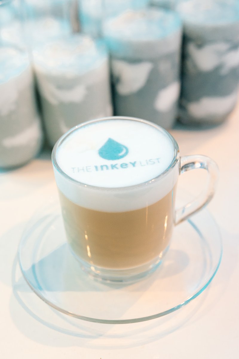 A clear glass mug holds a coffee latte with custom branding, featuring a blue droplet logo and 'THE INKEY LIST' text on 