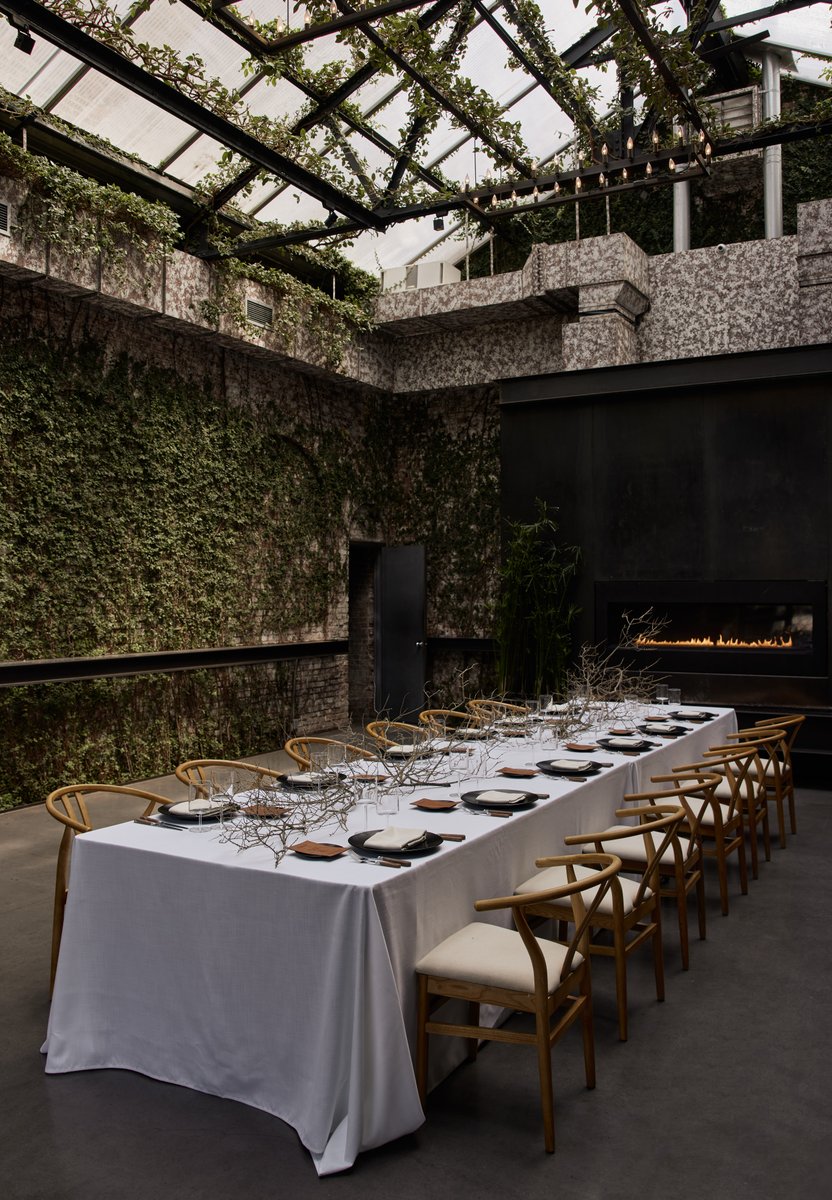 A long dining table is elegantly set in a unique venue, featuring ivy-covered brick walls and a glass ceiling that sugge