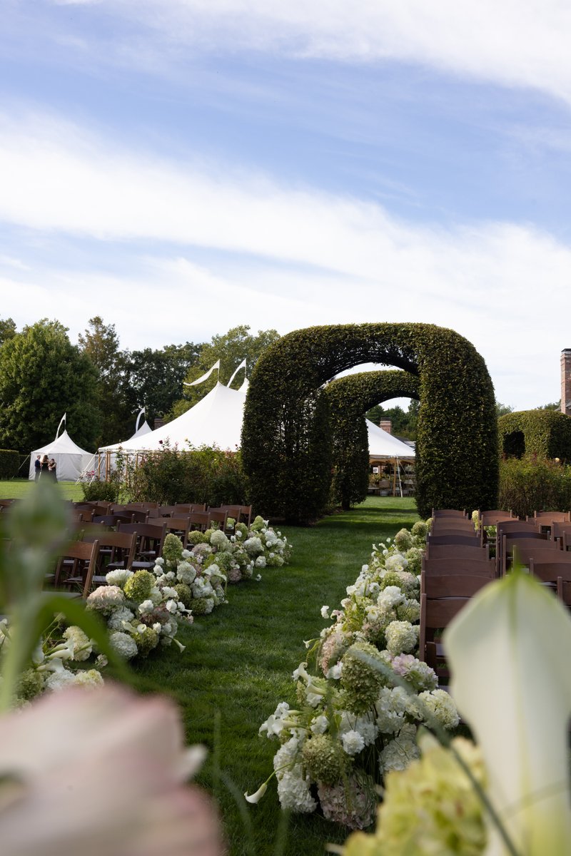 This image captures an elegant outdoor wedding ceremony aisle, lined with rows of wooden chairs and lush white and green