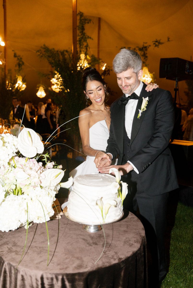A smiling bride and groom are captured in a joyful moment as they cut their wedding cake. The scene is set at an evening