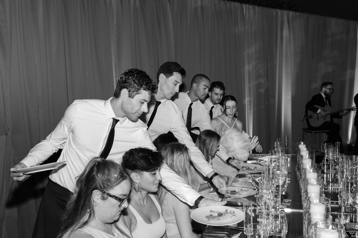 Servers in crisp white shirts and black ties are attentively serving plated dishes to guests seated at a long, elegantly