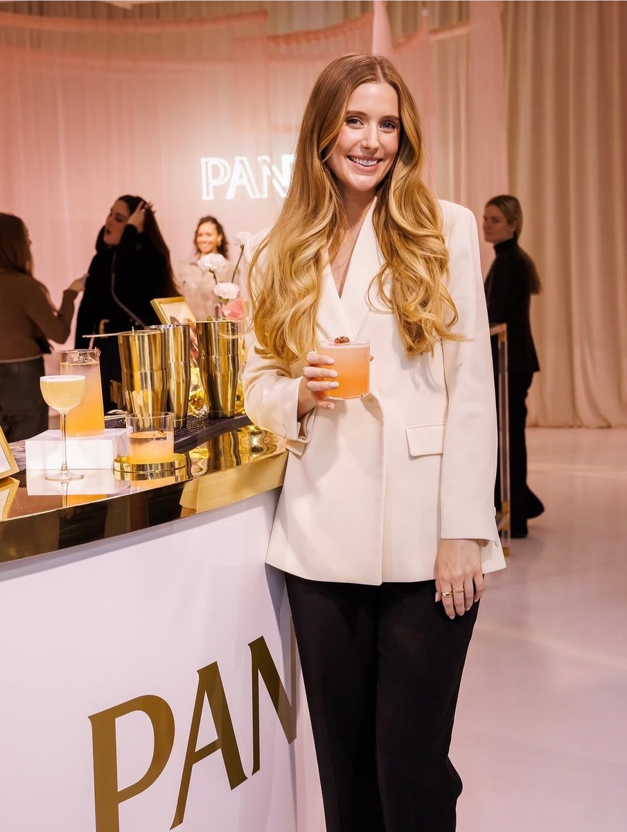 A smiling woman in a cream blazer holds a pink cocktail, leaning against a white and gold branded bar. In the soft pink-