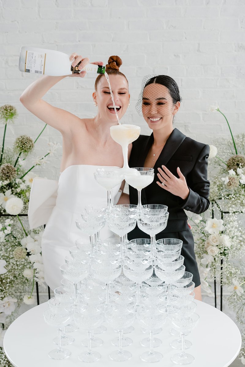 Two women are joyfully pouring champagne into a tall pyramid of coupe glasses, creating a festive champagne tower. One w