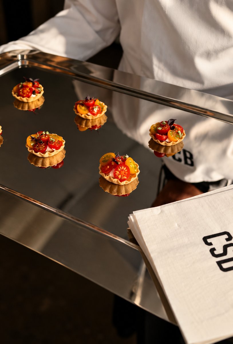A server in a white uniform holds a reflective silver tray presenting several colorful mini tarts topped with cherry tom