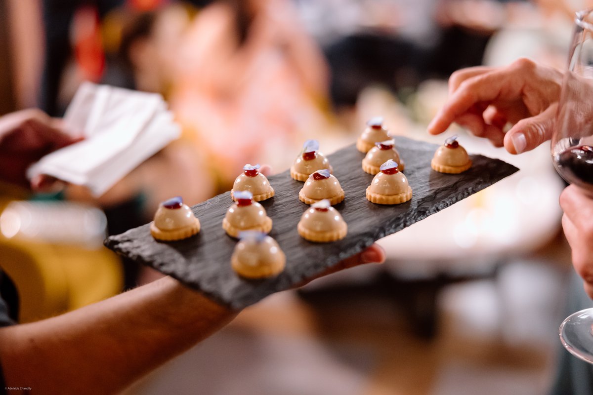 A server presents a slate tray of elegant, dome-shaped mini desserts to a guest. The guest's hand reaches for a dessert 