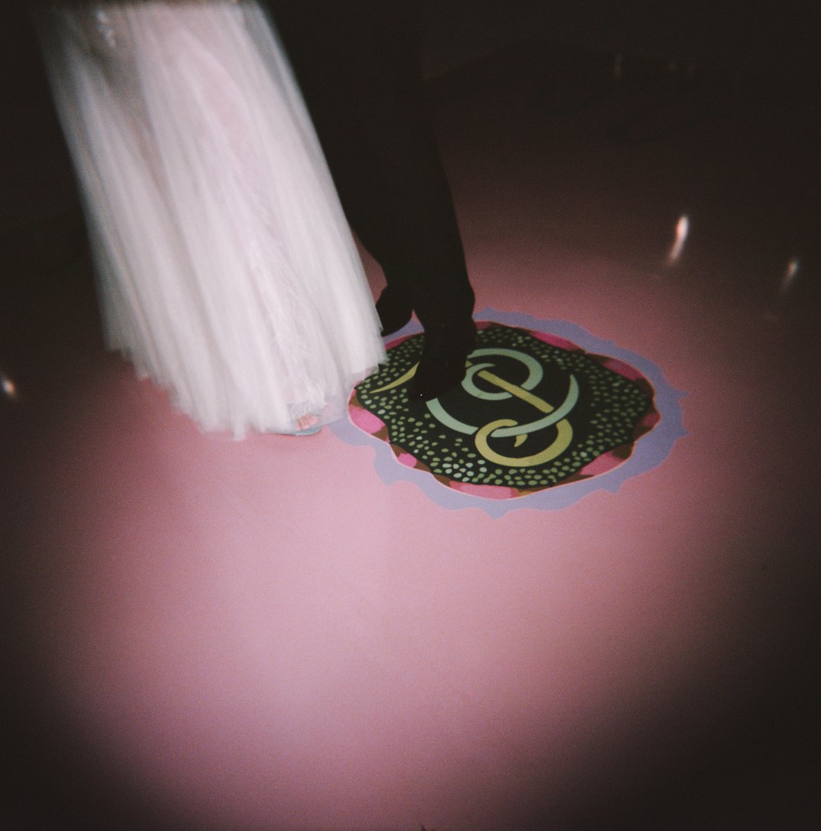 Two individuals, one in a flowing white dress and another in dark attire, stand on a vibrant pink floor. A colorful, abs