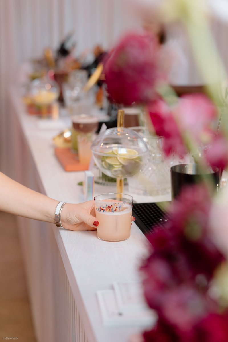 A close-up shot features a hand holding a light pink cocktail with a decorative floral rim, resting on a sleek white bar