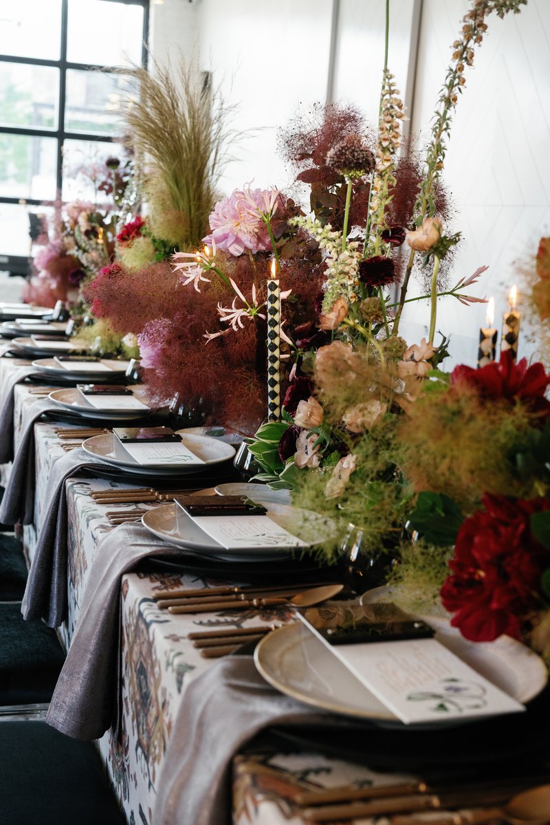 A long dining table is elegantly set for an event, featuring dramatic and lush floral arrangements in deep red, pink, an