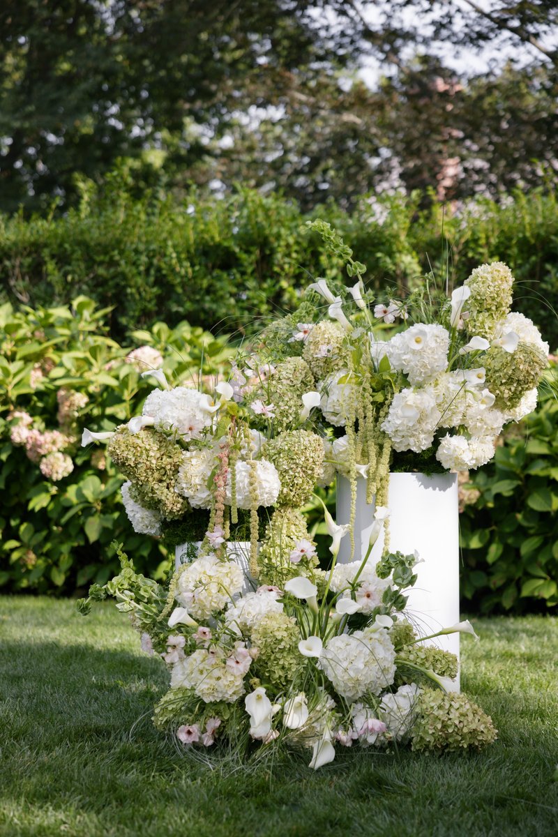 A large, elegant floral arrangement featuring white hydrangeas, calla lilies, and green foliage is displayed outdoors. T