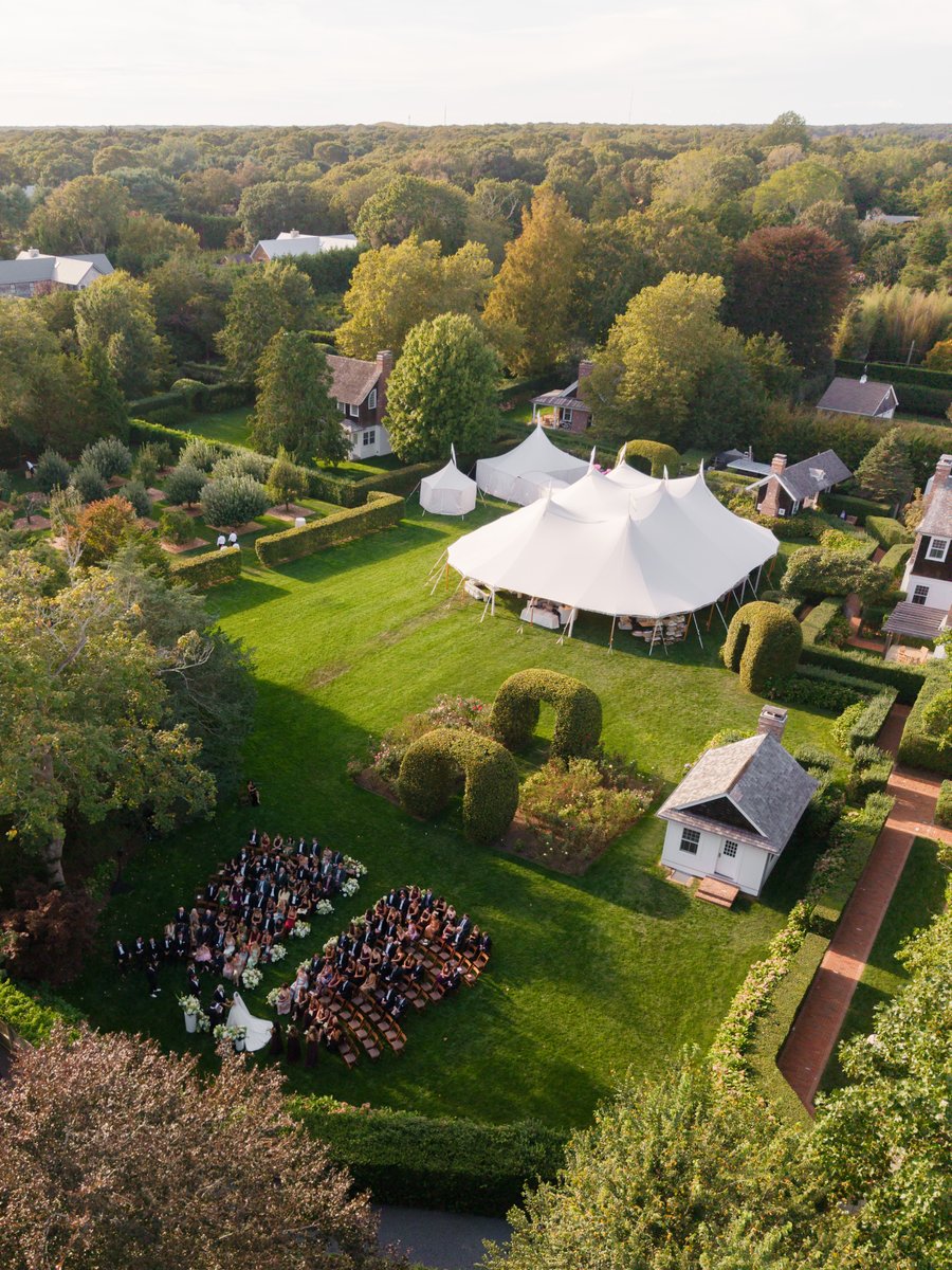 An aerial view captures an elegant outdoor wedding ceremony in a lush garden setting. Guests are seated on chairs facing