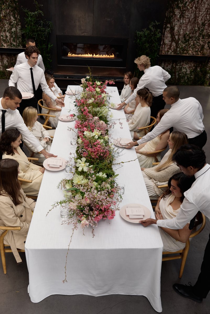 Servers in white shirts and black ties are presenting place settings to guests seated at a long, elegantly decorated tab