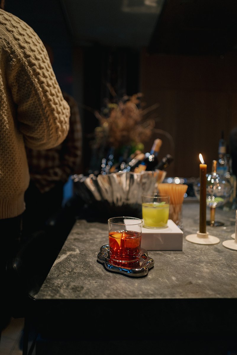 A close-up shot of two cocktails on a dark stone bar, illuminated by a single candle. A person in a textured sweater is 