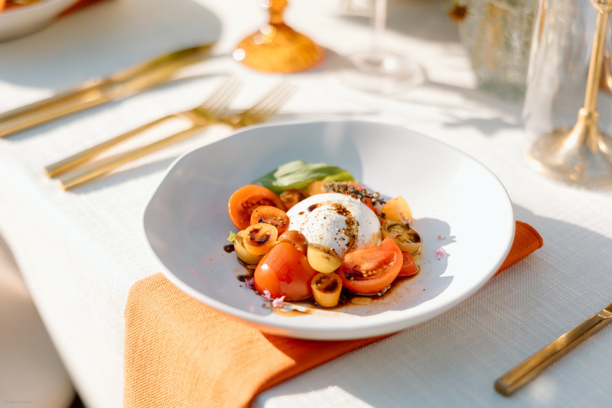 A vibrant close-up of a burrata and tomato salad, artfully arranged in a white bowl on a crisp white tablecloth. Gold cu