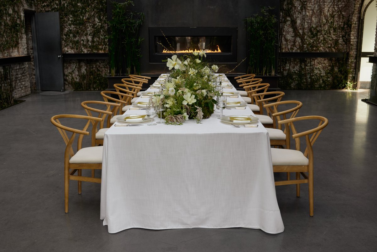 A beautifully set long dining table awaits guests in a modern event space. The table is adorned with a lush, organic flo
