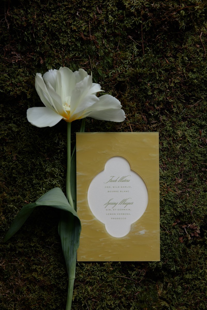 A delicate white tulip and a stylish yellow-framed menu card are artfully arranged on a bed of dark green moss. The menu