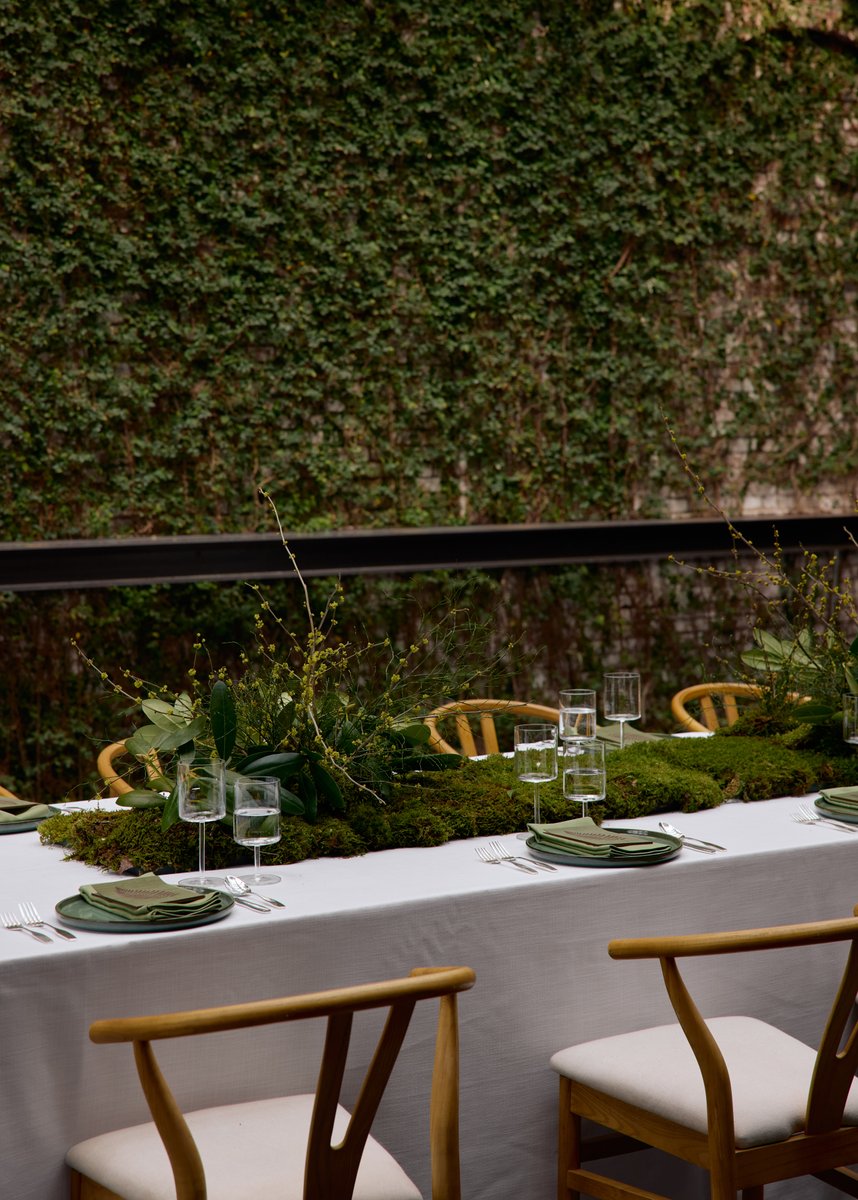 A beautifully set long table features a lush, natural centerpiece made of moss and various green foliage, creating an or