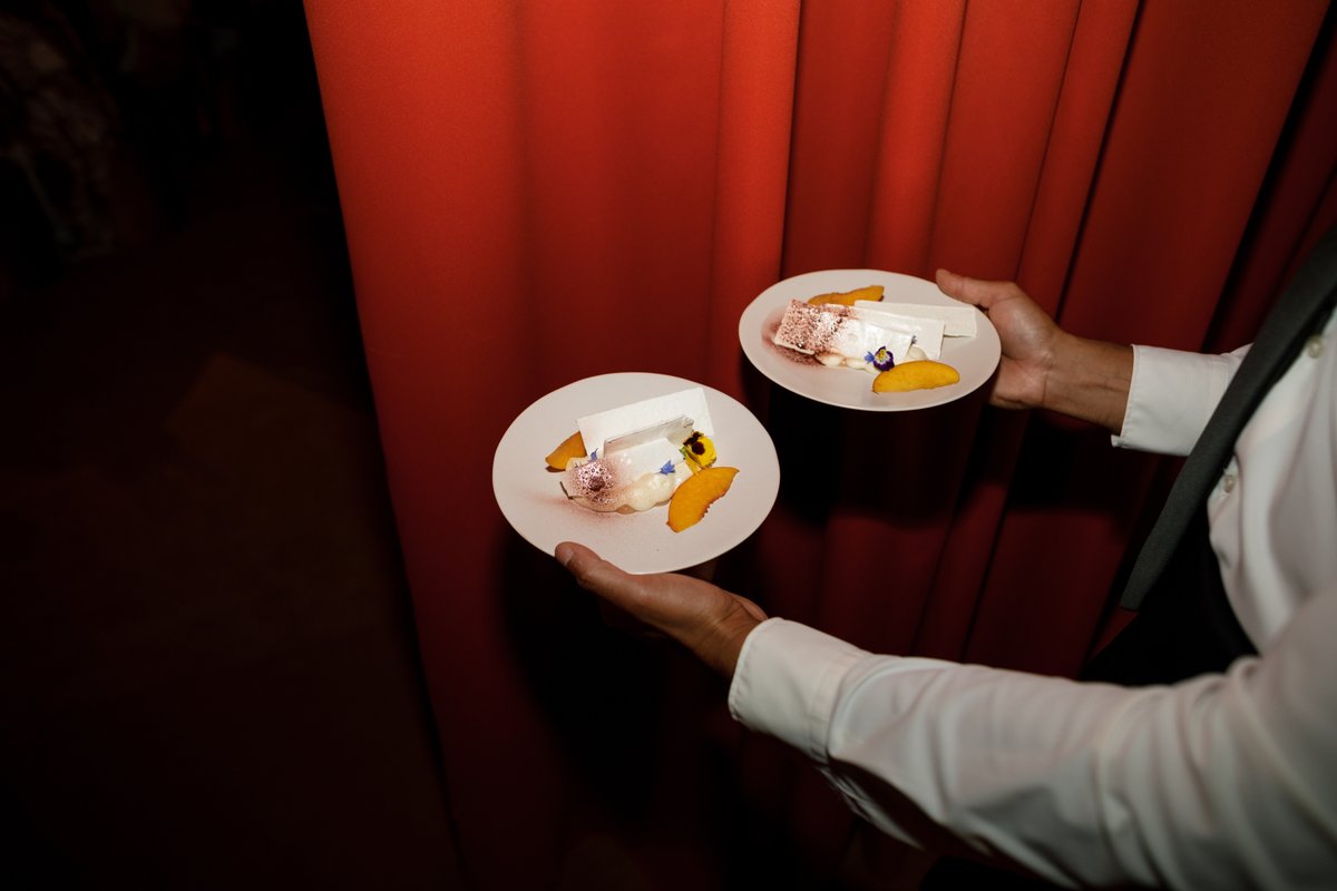 A server, dressed in a white shirt and dark tie, holds two elegantly plated desserts against a rich red curtain backdrop