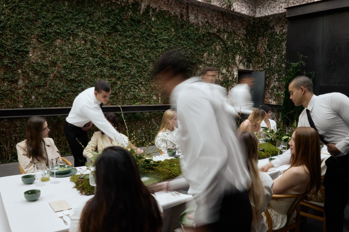 This dynamic medium-shot captures servers attending to guests seated at a long table in a venue with a striking ivy-cove