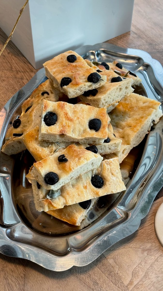 A close-up shot features numerous pieces of freshly baked focaccia bread, topped with black olives, arranged on a decora