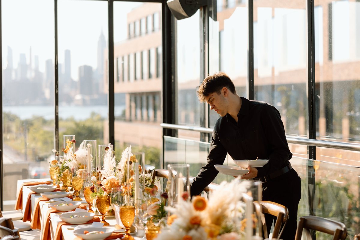 A server in a black uniform meticulously sets a long dining table adorned with elaborate floral arrangements and amber g