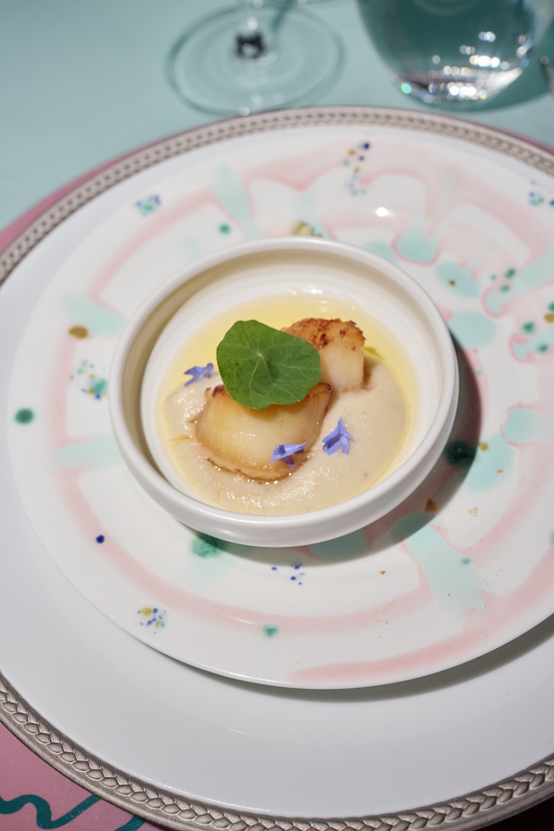 A beautifully plated appetizer features two seared scallops atop a creamy puree, garnished with a vibrant green leaf and