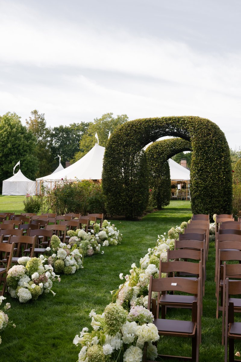 An elegant outdoor ceremony space is prepared on a lush green lawn. Rows of dark wooden chairs flank an aisle adorned wi
