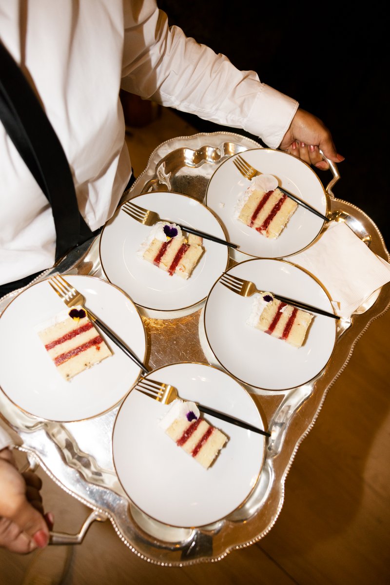 A server in a white shirt and black suspenders carries a silver tray laden with several slices of cake. Each slice is el