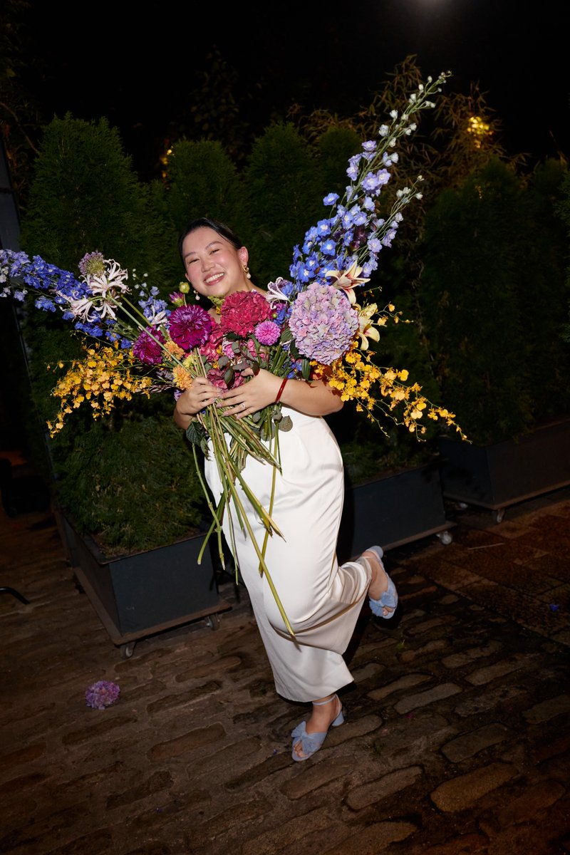 A smiling woman in white pants and blue shoes joyfully holds a large, vibrant bouquet of mixed flowers, including purple