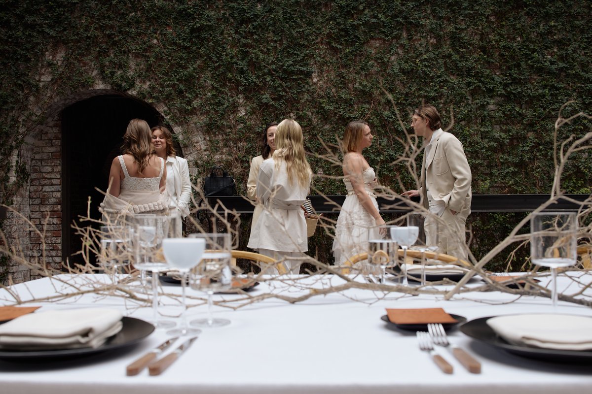 A group of guests, dressed in light-colored attire, are socializing in an outdoor setting with an ivy-covered brick wall
