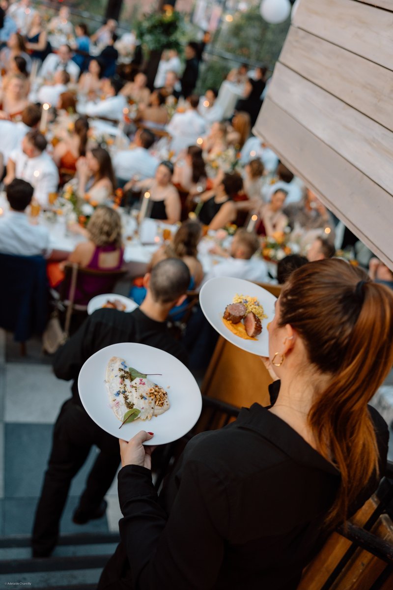 A server, seen from behind, carries two elegantly plated dishes down a staircase, with a bustling event of guests dining