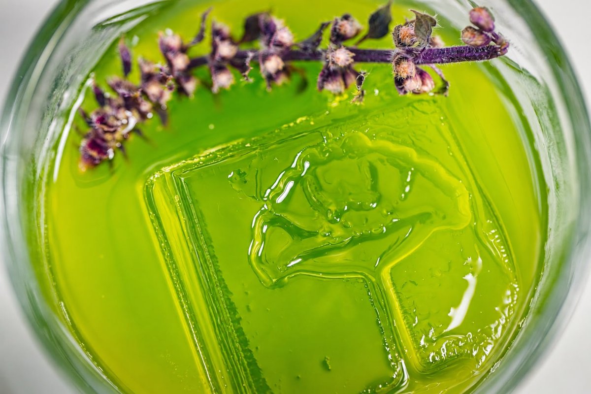 A vibrant green cocktail with a large square ice cube is garnished with a sprig of purple basil, viewed from directly ab