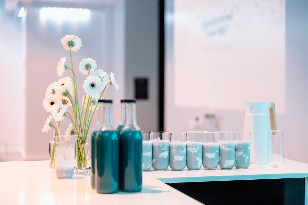 A modern beverage station is set up with several glasses containing a frothy light blue drink, alongside bottles of a da