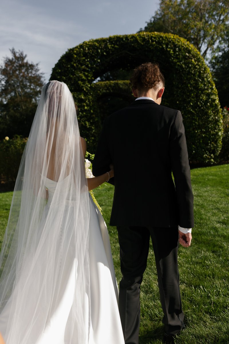 A couple, likely a bride and groom, walks away from the camera through a sunny garden. The bride wears a white dress and