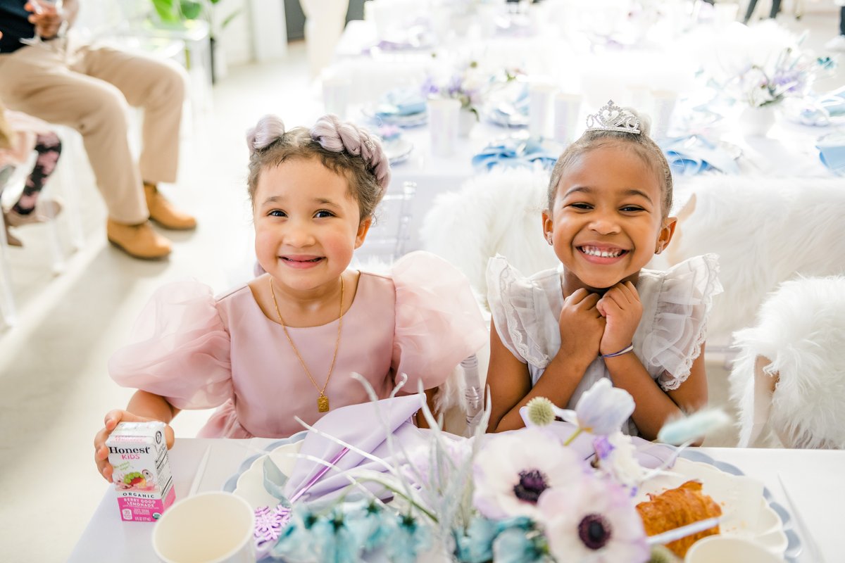 Two young girls, dressed in festive attire, are seated at a beautifully decorated table and smiling brightly at the came
