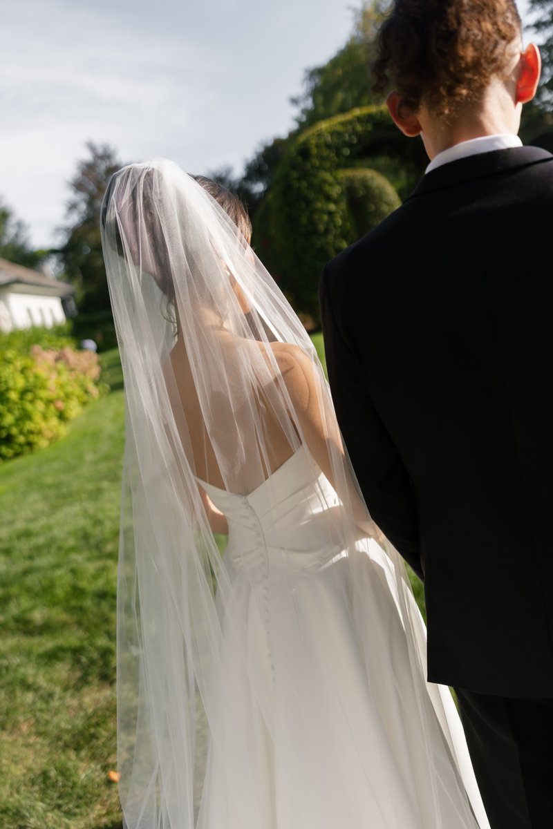 A bride in a white gown and veil walks alongside a groom in a black suit, seen from behind. They are walking on a grassy