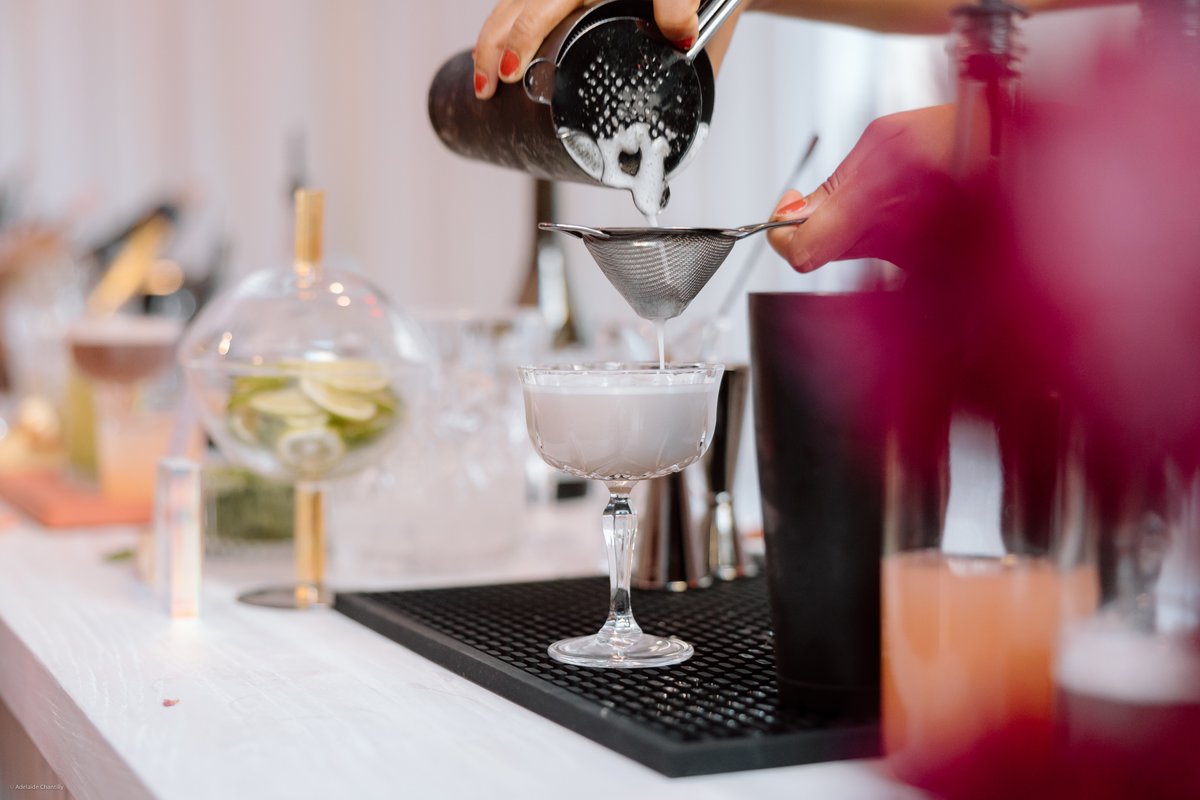 A close-up shot captures a bartender's hands expertly straining a creamy white cocktail from a shaker into an elegant co