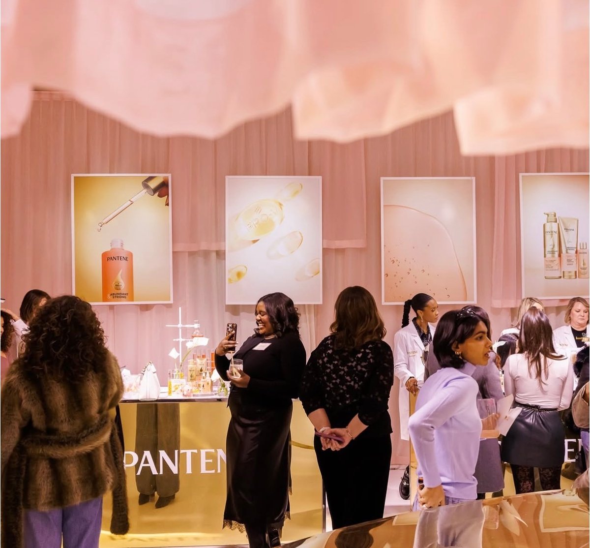 Guests mingle around a gold-branded Pantene station at an indoor event. One guest smiles while taking a photo with her p