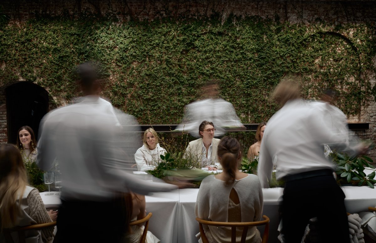 Servers, blurred by motion, attend to guests seated at a long table adorned with lush green decor. The backdrop is a str