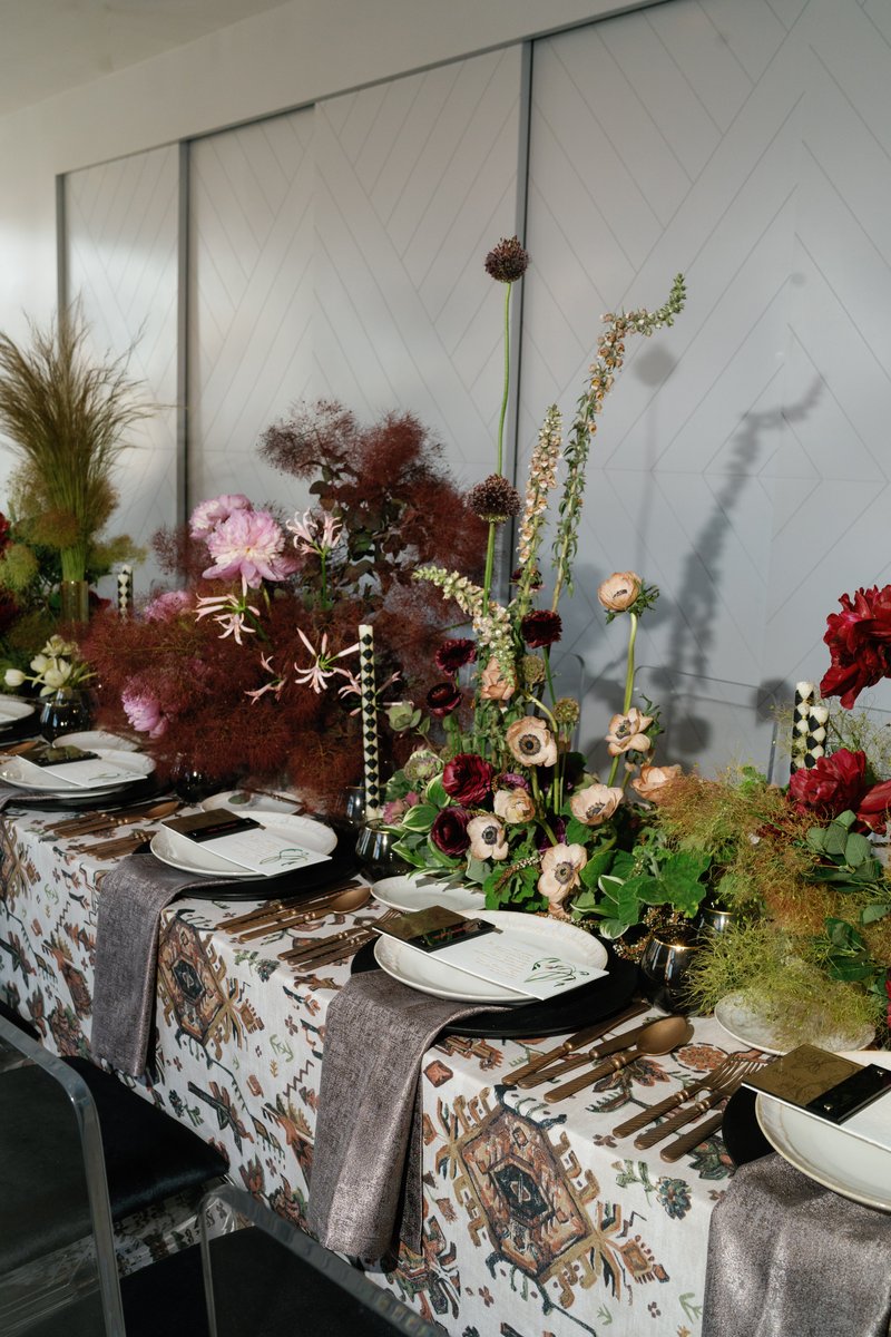 A beautifully arranged long table is set for an event, featuring an eclectic mix of lush floral arrangements in shades o