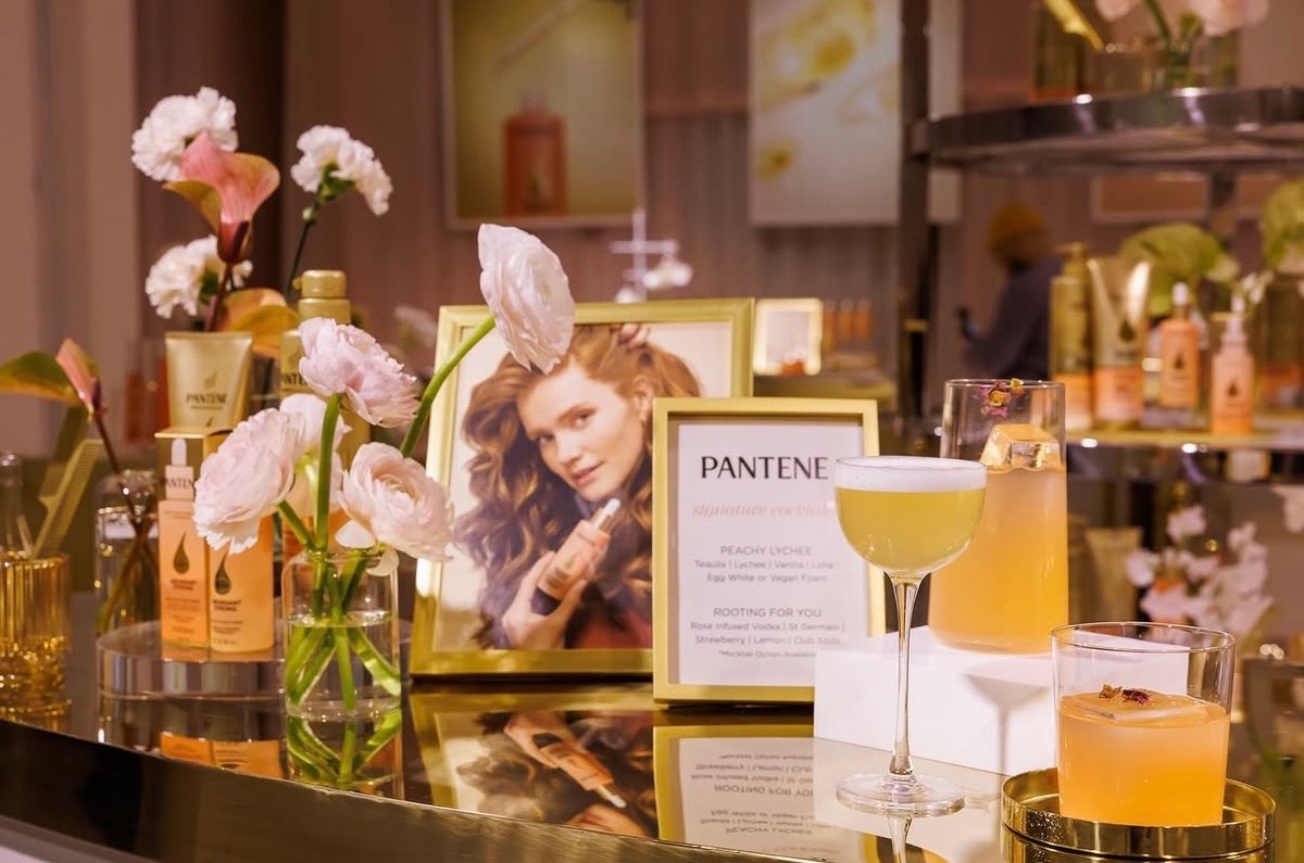 A stylish cocktail station is set up, featuring three signature drinks alongside prominent Pantene branding. The display