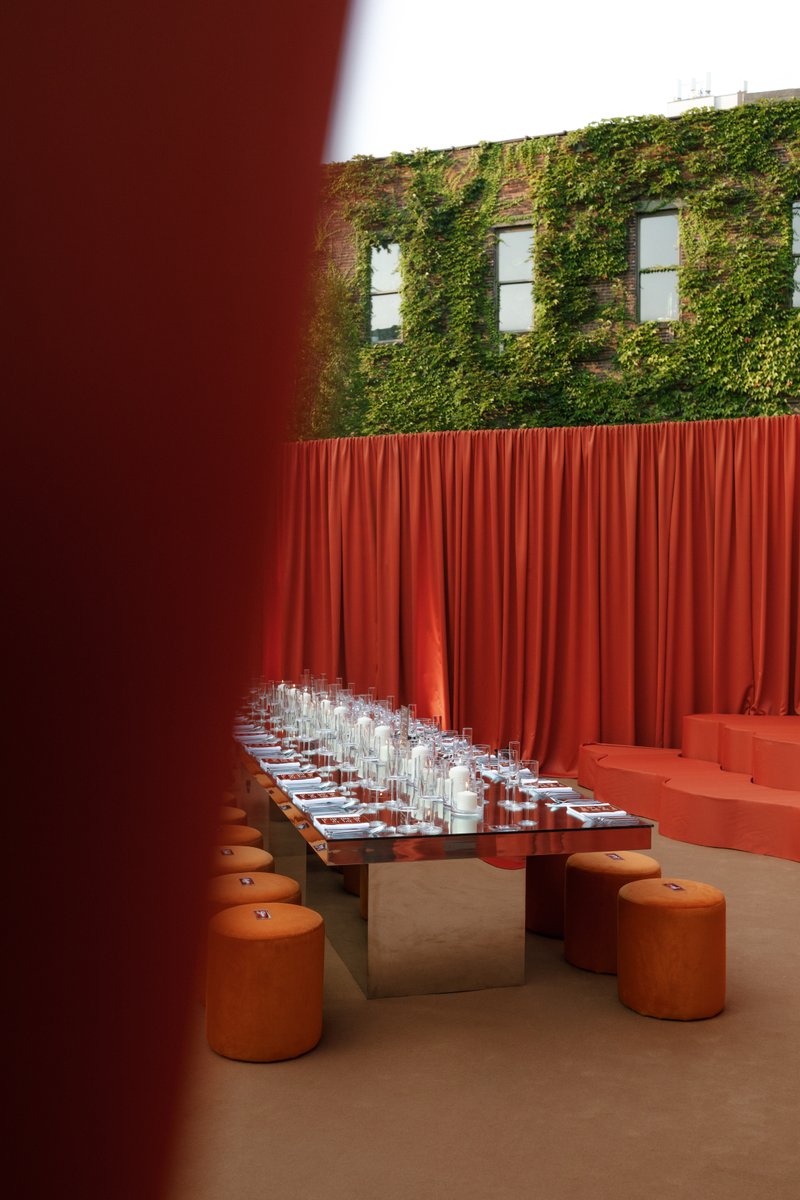 A long, mirrored dining table is meticulously set with glassware and white candles, flanked by orange velvet stools, aga