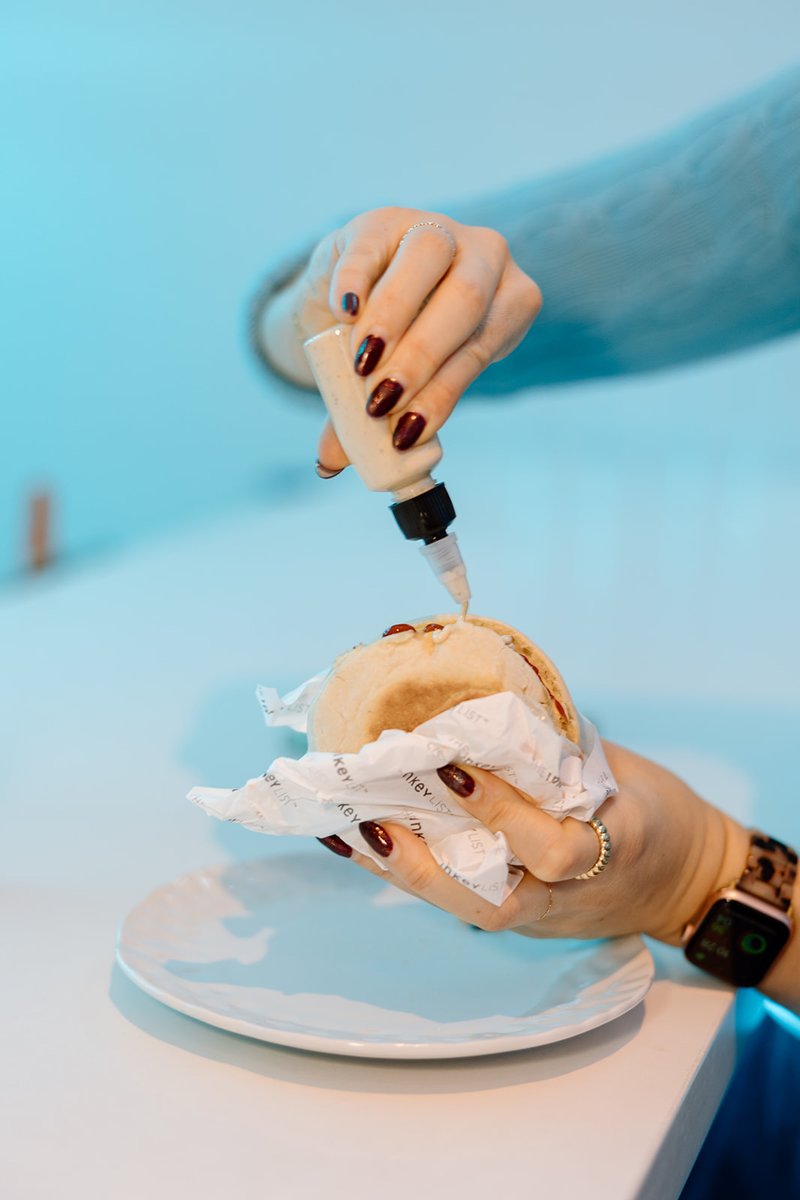 A close-up shot shows hands adding a creamy sauce from a squeeze bottle onto a burger or sandwich wrapped in branded pap