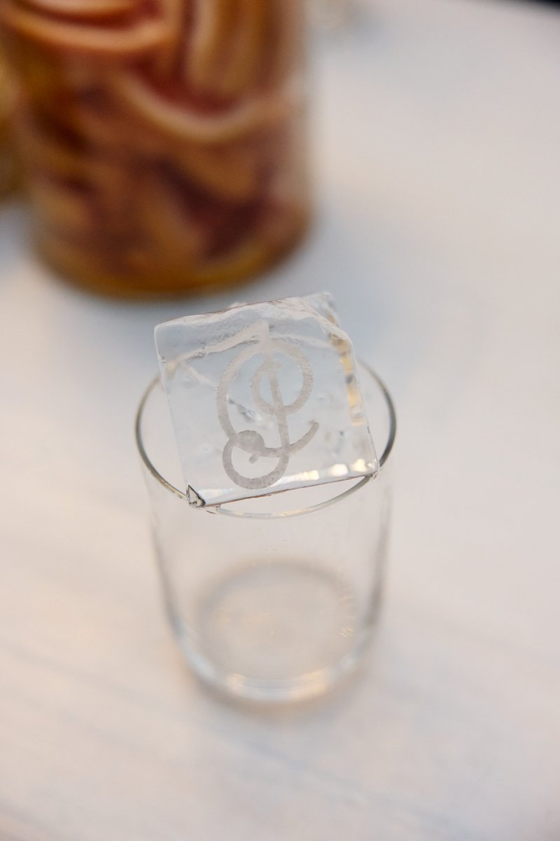 A clear glass holds a large, custom ice cube featuring a monogrammed 'AD' or 'AP' design. In the softly blurred backgrou