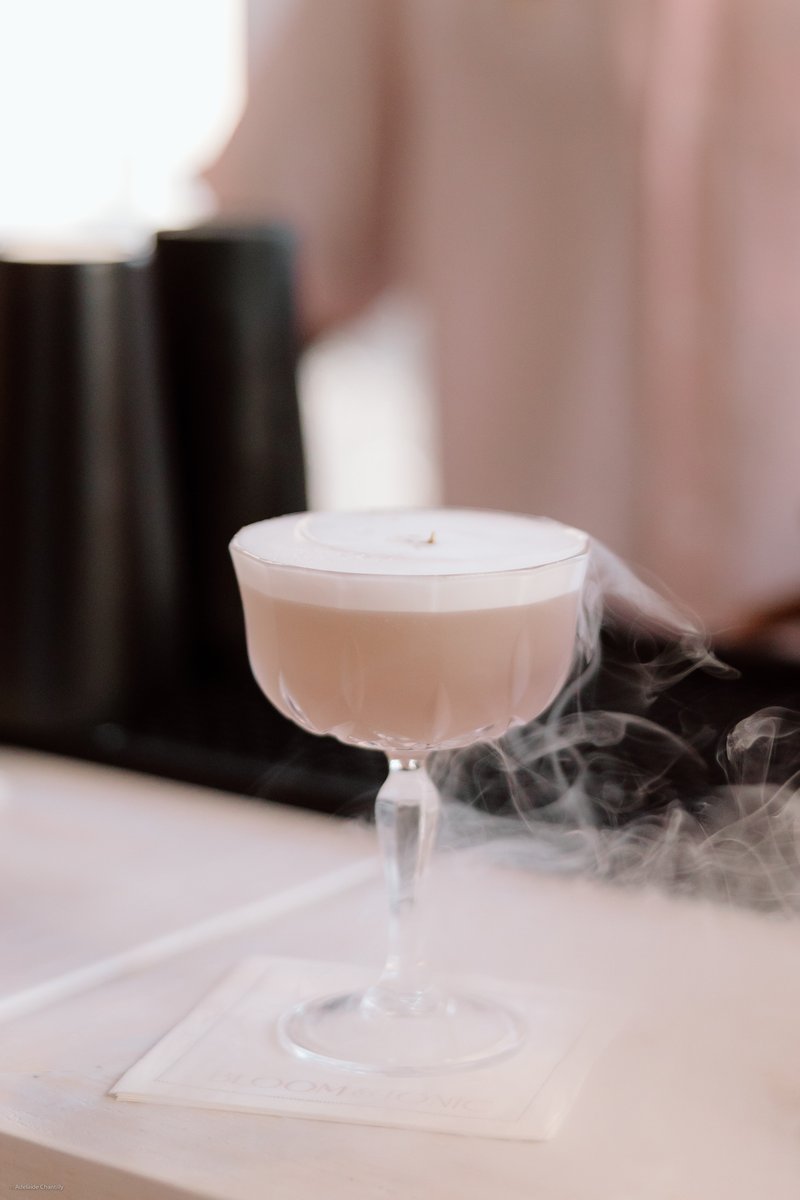 A beautifully presented cocktail in a coupe glass, featuring a frothy top and an intriguing smoke effect emanating from 