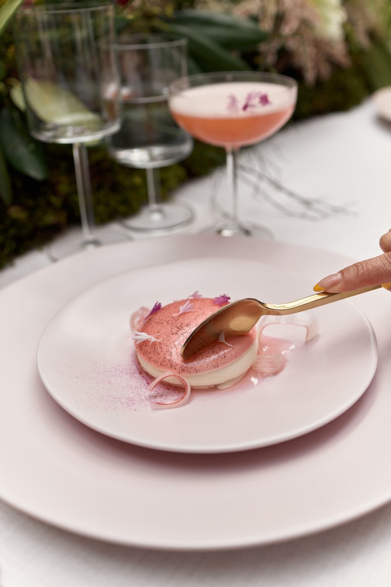 A close-up shot captures a hand with a golden spoon about to scoop into an elegant pink and white layered dessert, artfu
