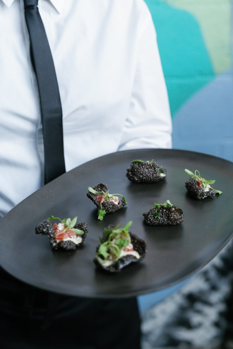 A server, dressed in a crisp white shirt and black tie, holds a sleek black oval tray presenting several artfully arrang