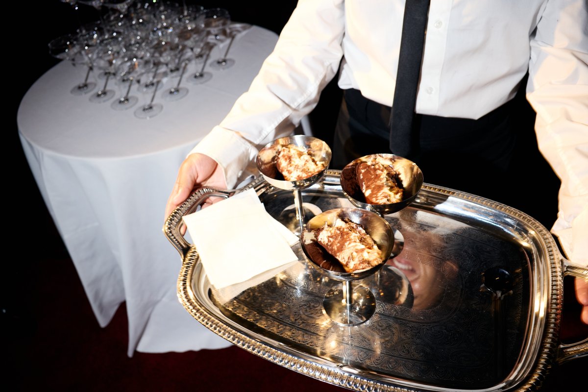 A server in a white shirt and black tie holds a silver tray with three individual dessert servings. In the background, a