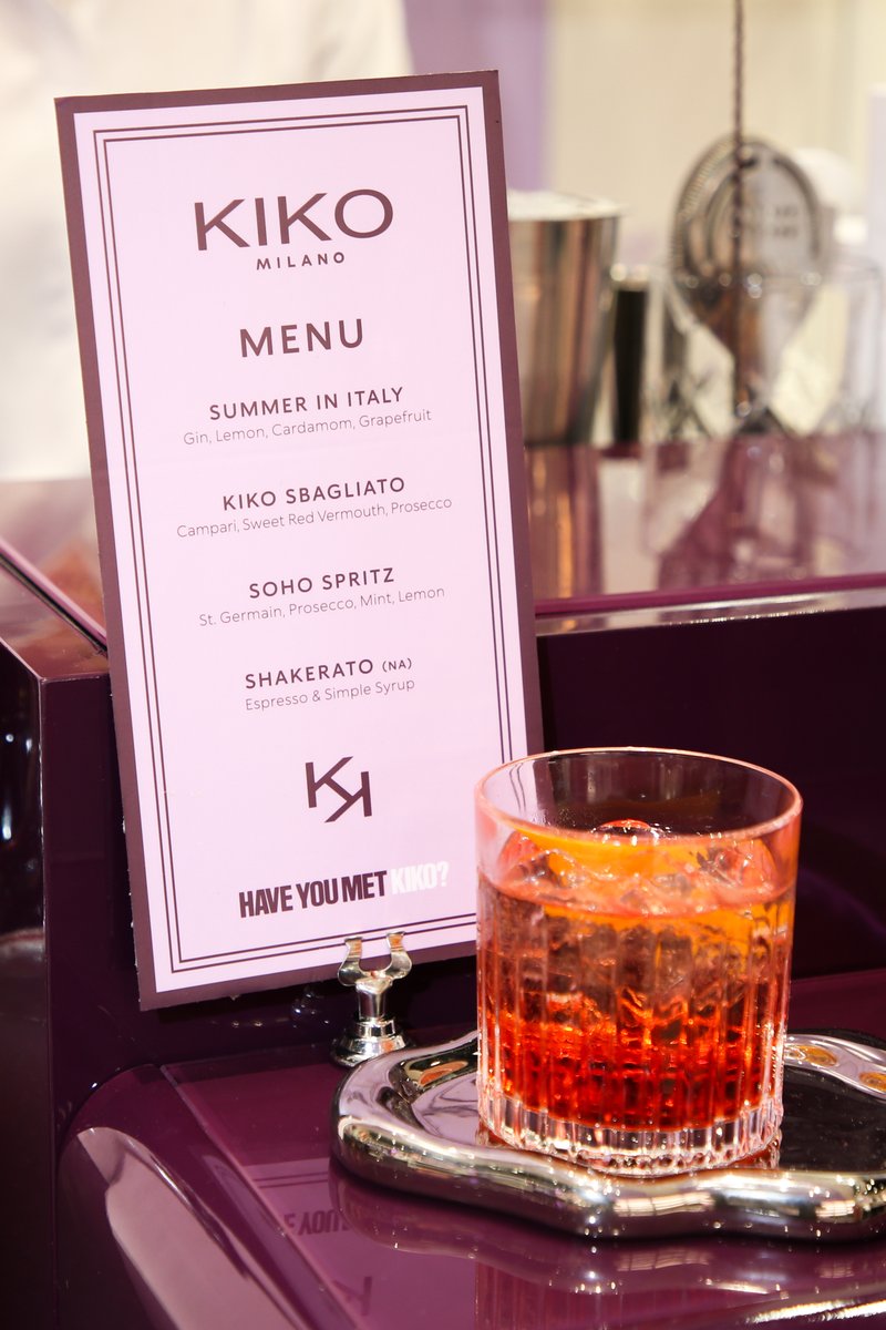 A branded cocktail menu for Kiko Milano stands prominently next to a vibrant red-orange cocktail, served in a rocks glas