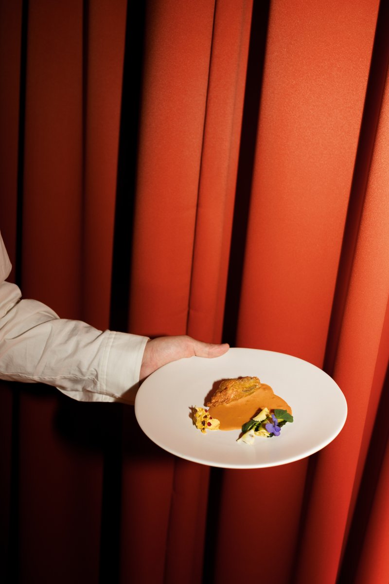 A server's arm, dressed in a white shirt, holds a white plate featuring a meticulously plated dish against a dramatic re