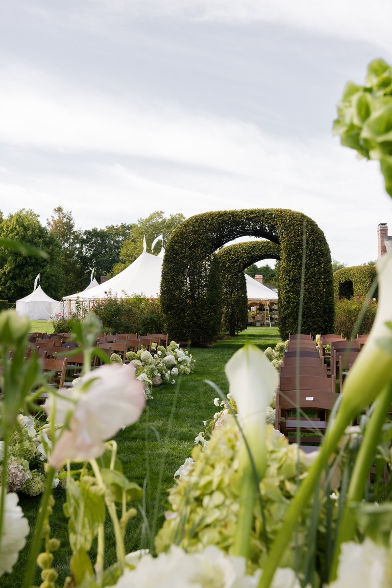 An elegant outdoor wedding ceremony aisle is set up with rows of wooden chairs on either side of a lush green lawn. The 
